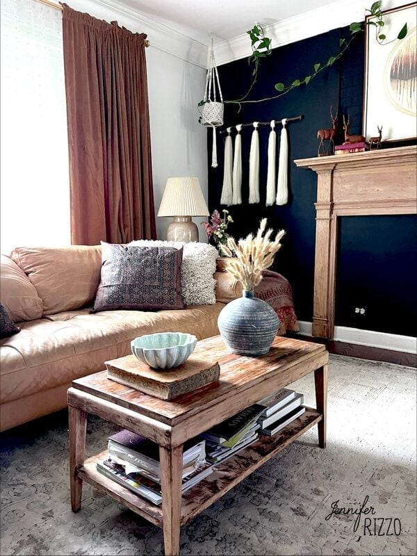 A cozy living room with a tan leather sofa, wooden coffee table with books and decor, dark accent wall, and pampas grass in a vase.