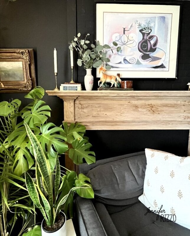 Living room corner with a gray armchair, potted plants, a wooden mantel decorated with vases, candlesticks, a horse figurine, and abstract art on a dark wall.