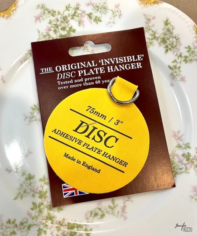 A yellow adhesive plate hanger labeled "DISC" is attached to its packaging, which reads "The Original 'Invisible' DISC Plate Hanger, 75mm / 3", Made in England," next to a floral plate.