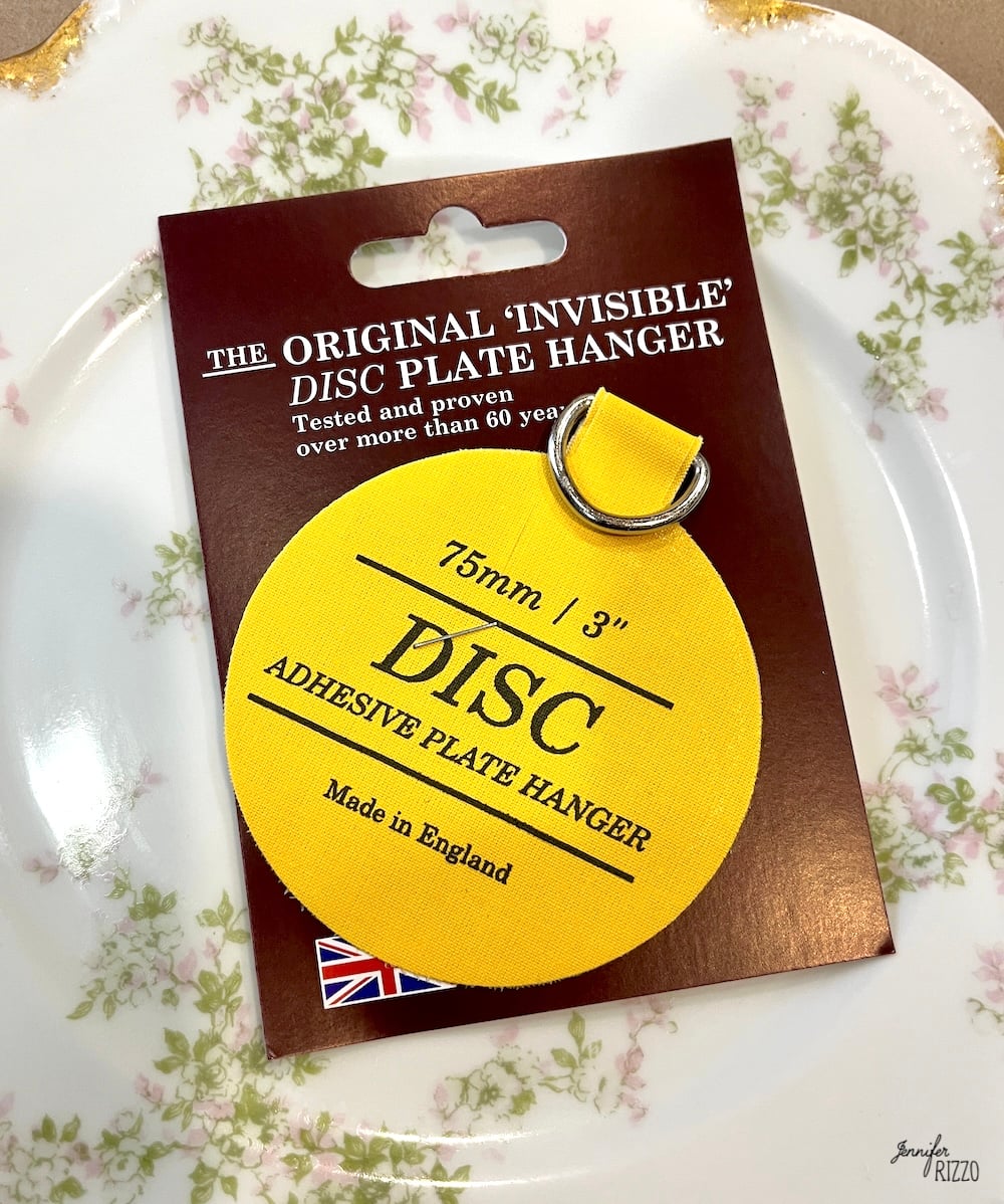 A yellow adhesive plate hanger labeled "DISC" is attached to its packaging, which reads "The Original 'Invisible' DISC Plate Hanger, 75mm / 3", Made in England," next to a floral plate.