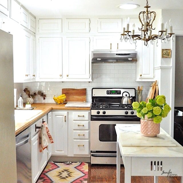 A bright kitchen with white cabinets, stainless steel stove and dishwasher, wooden countertops, a small island, and a chandelier. A vase of green flowers sits on the island.