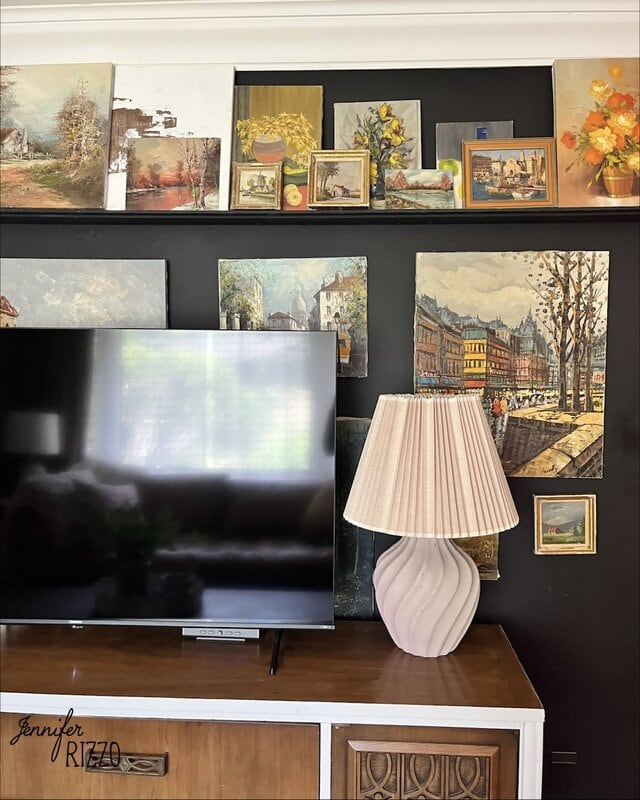 A flat-screen TV, a white lamp with a pleated shade, and a wooden cabinet are placed in front of a black wall decorated with framed artwork and paintings.