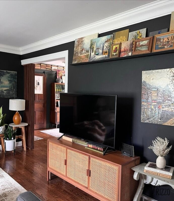 A living room with dark walls, a TV on a wooden cabinet, artwork displayed above, a plant, and a glimpse into an adjoining room with wooden flooring.
