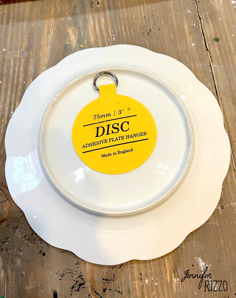 A white plate is shown upside down with a yellow adhesive plate hanger labeled "75mm | 3'' DISC Adhesive Plate Hanger Made in England" attached to its back.
