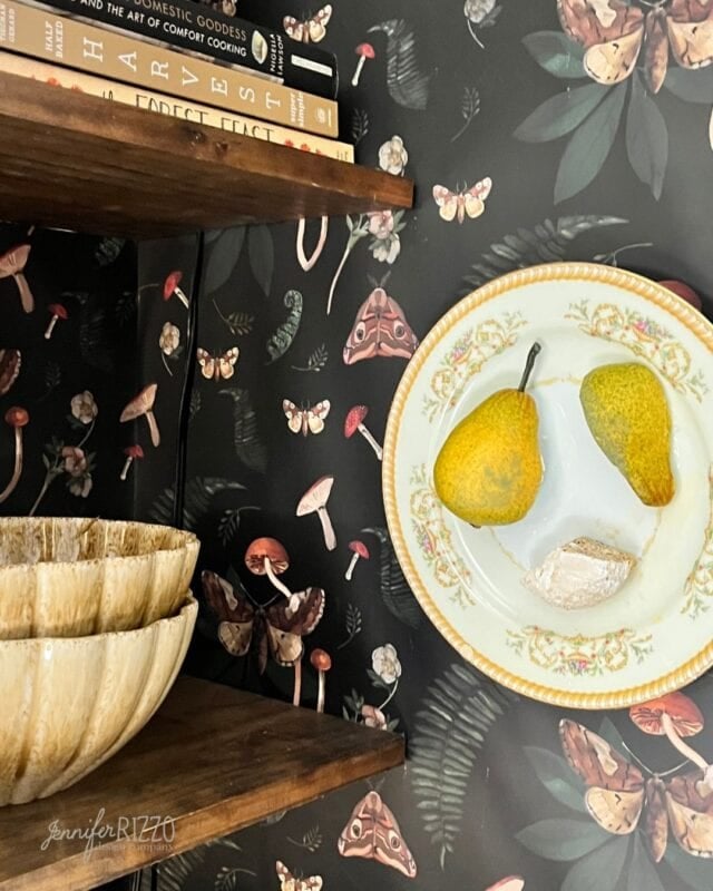 A decorative plate with two pears and a small rock sits on a shelf next to stacked bowls; books and a floral-mushroom patterned wallpaper are in the background.