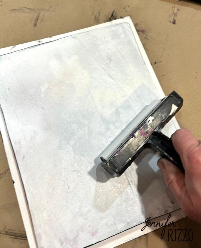A hand uses a brayer to spread white paint over a square canvas on a brown surface.