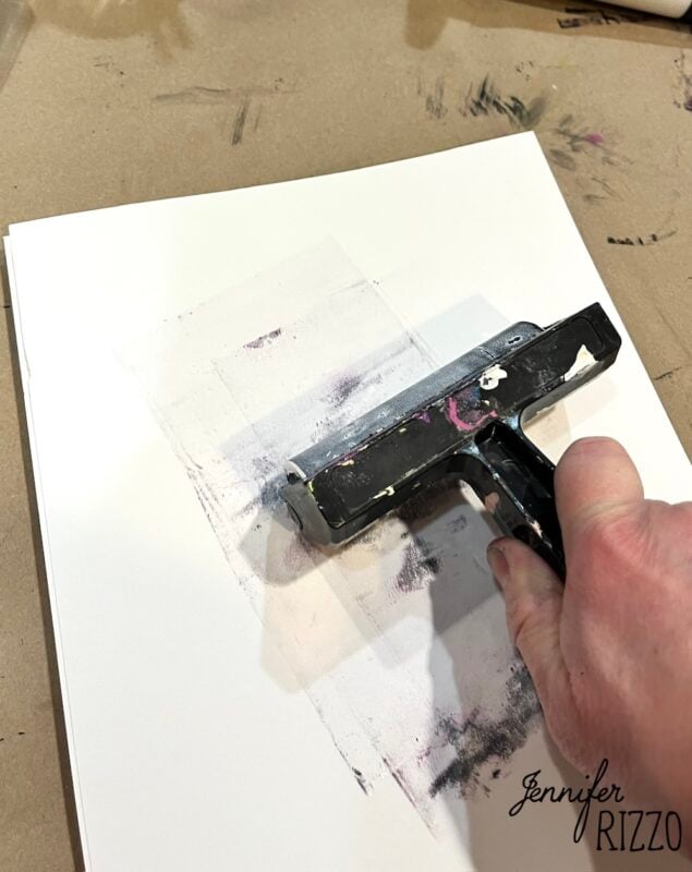 A hand uses a brayer to roll ink or paint onto a sheet of paper on a work surface, with the name "Jennifer Rizzo" written in the corner.