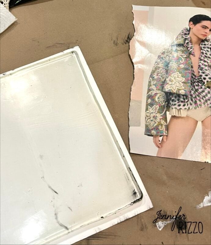 A torn fashion magazine page showing a model in an ornate jacket lies next to a white rectangular tray on a brown surface. "Jennifer Rizzo" is printed at the bottom right.