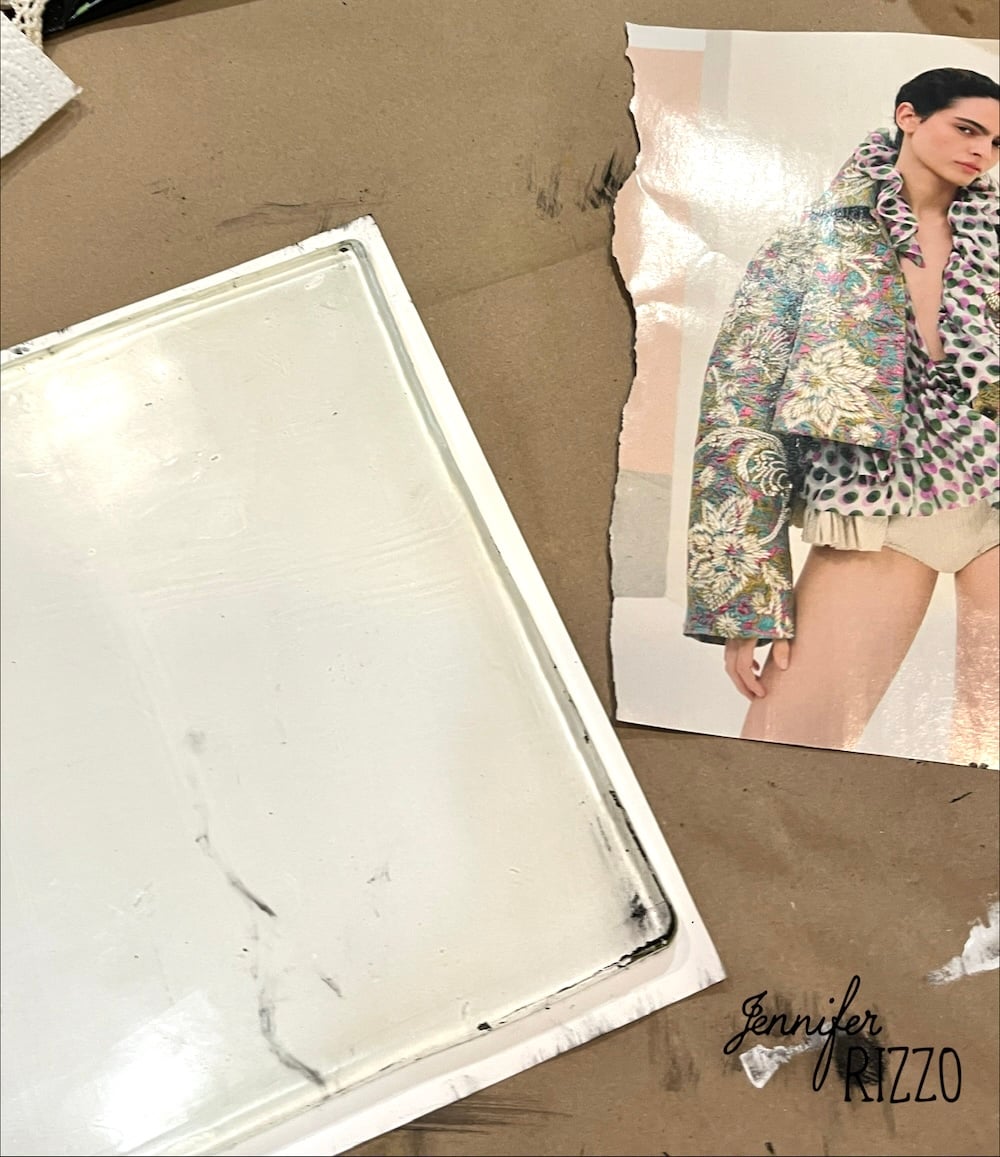 A torn fashion magazine page showing a model in an ornate jacket lies next to a white rectangular tray on a brown surface. "Jennifer Rizzo" is printed at the bottom right.