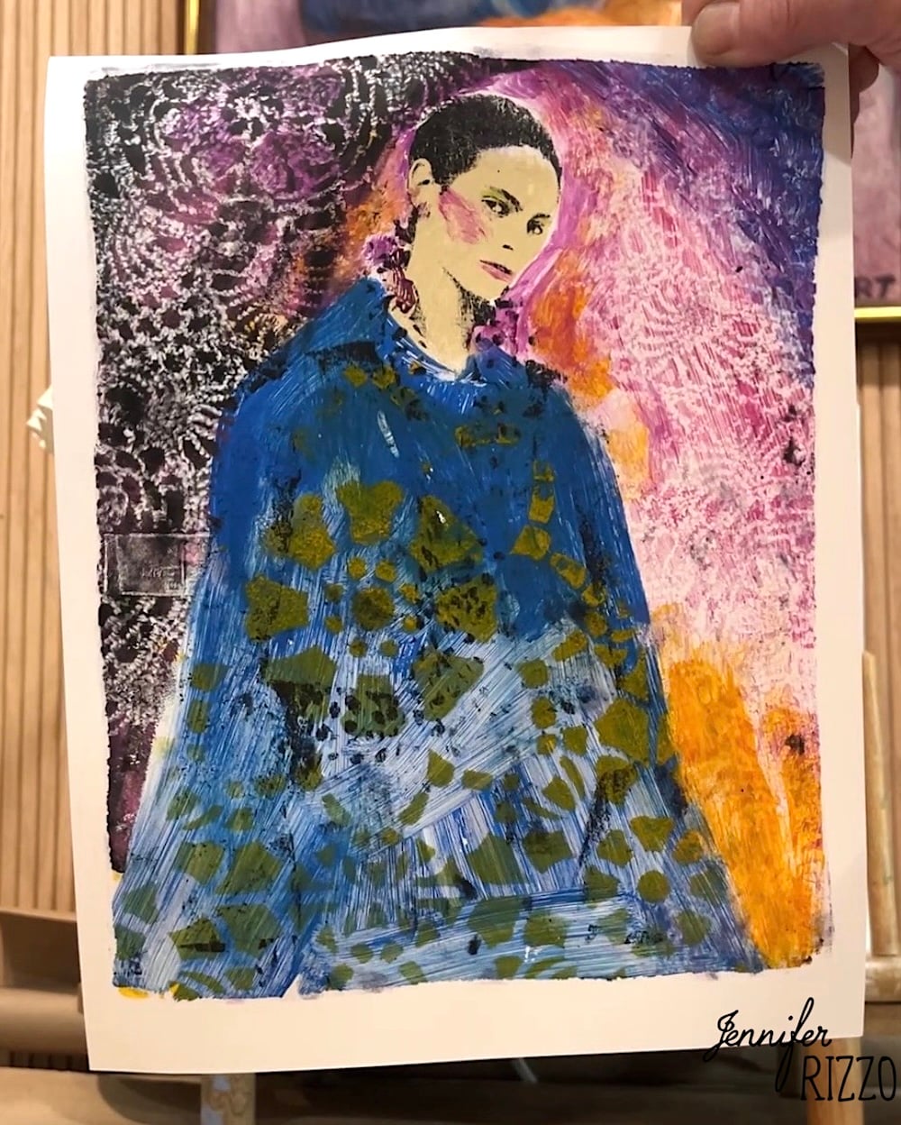 A hand holds a colorful portrait of a woman in a blue patterned garment against a vibrant, textured background. The image is signed "Jennifer Rizzo" in the corner.