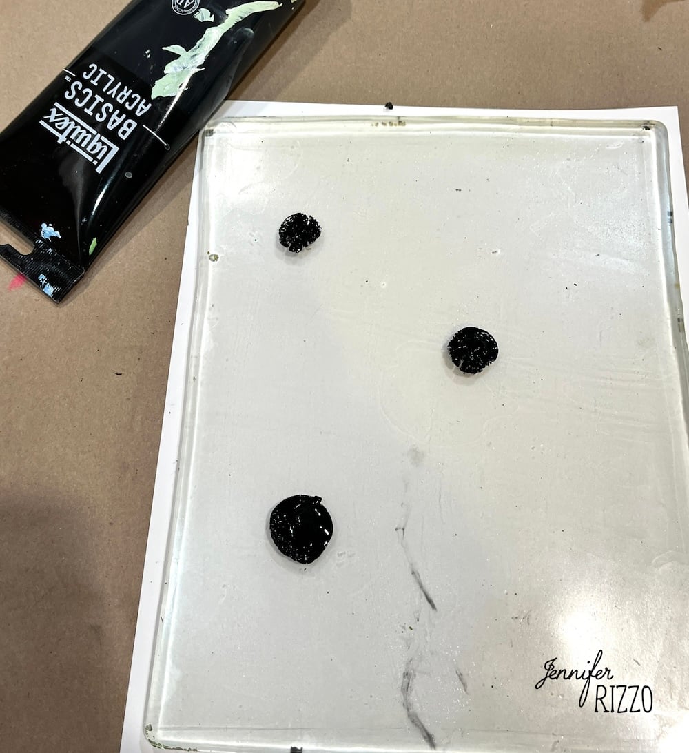 A glass palette with three blobs of black acrylic paint and a tube of Liquitex Basics acrylic paint on the side. The corner shows the watermark "Jennifer Rizzo.