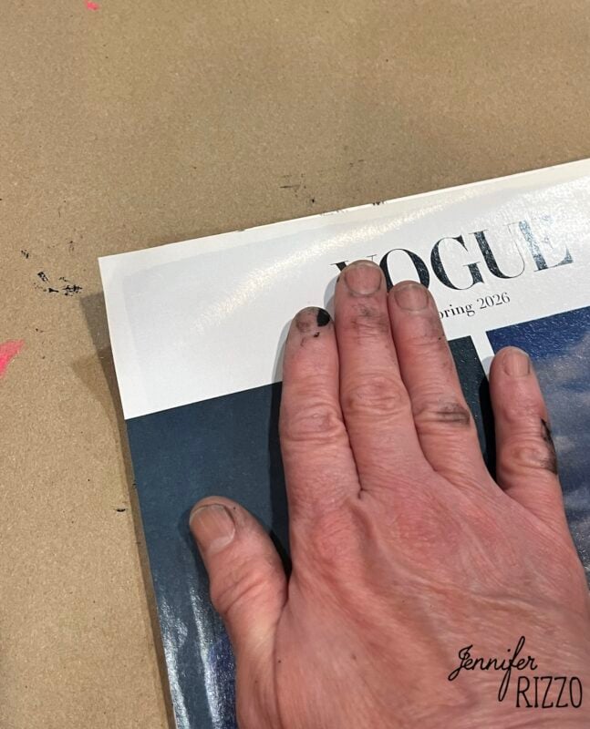 A hand with ink-stained fingers presses down on the corner of a magazine with a partially visible "VOGUE" title.