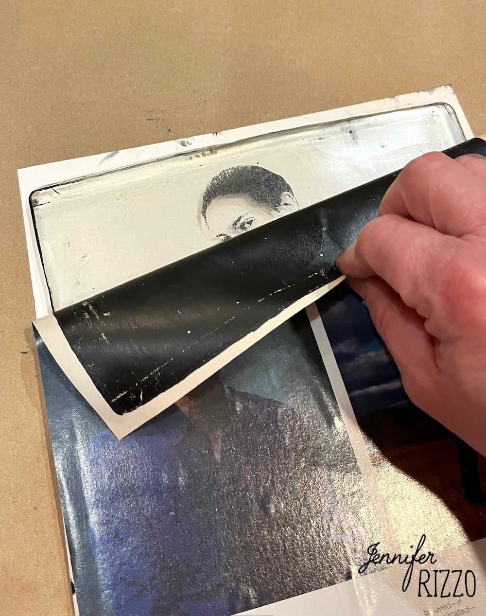 A hand peels back a sheet of photographic paper to reveal a black and white image transfer onto a canvas, with other photos beneath and the name Jennifer Rizzo in the corner.