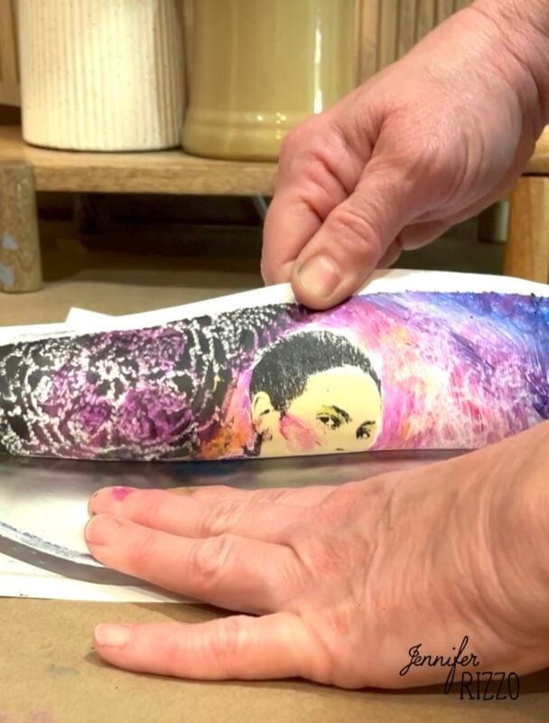 Close-up of hands transferring a printed image of a woman's face onto a curved surface, with art supplies and containers in the background.