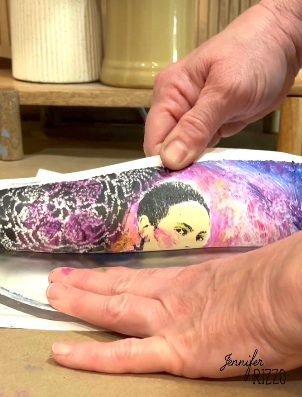 Close-up of hands transferring a printed image of a woman's face onto a curved surface, with art supplies and containers in the background.