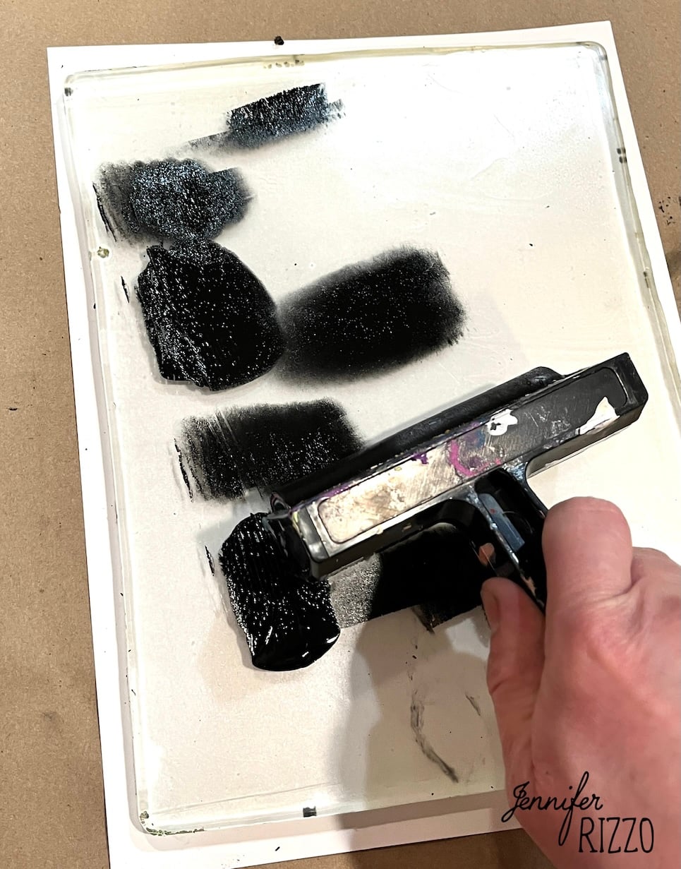 A hand uses a roller to spread black ink on a glass surface, with several black ink patches visible.