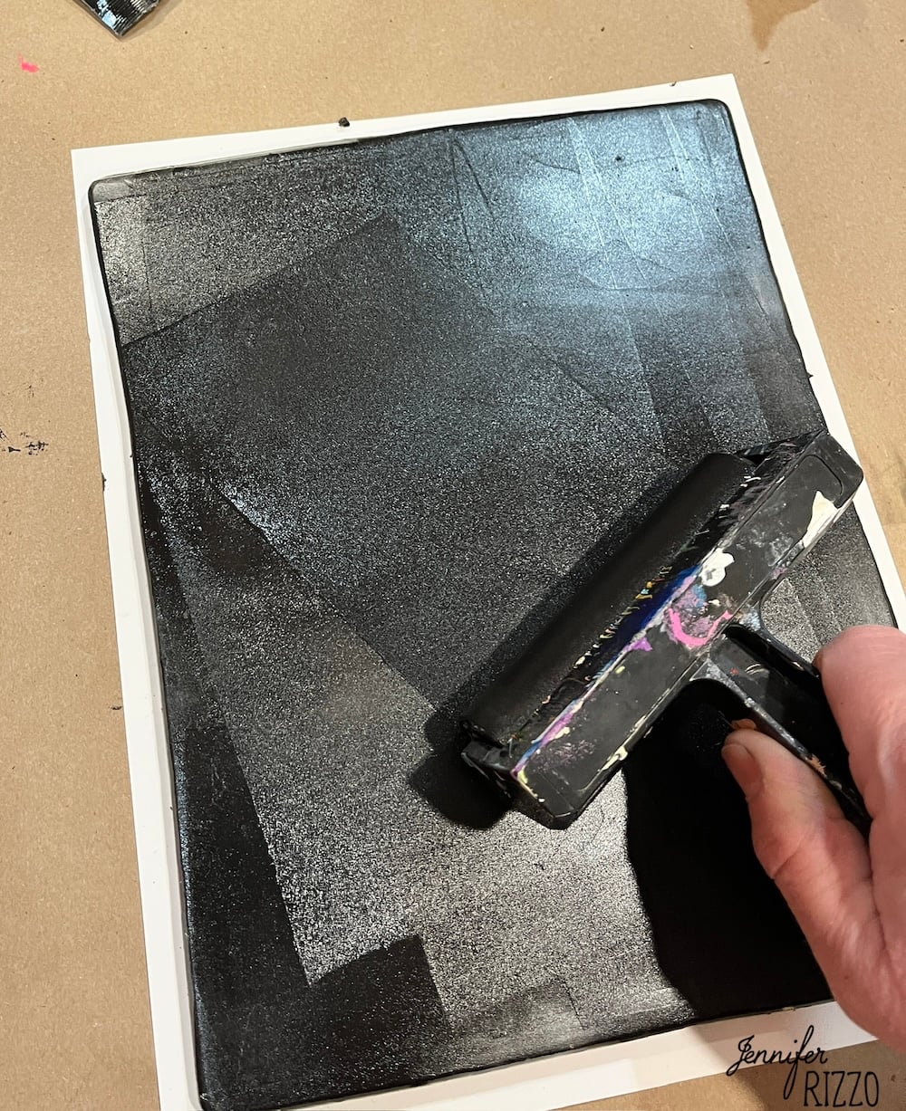 A hand uses a brayer to roll black ink onto a rectangular surface, covering it evenly.