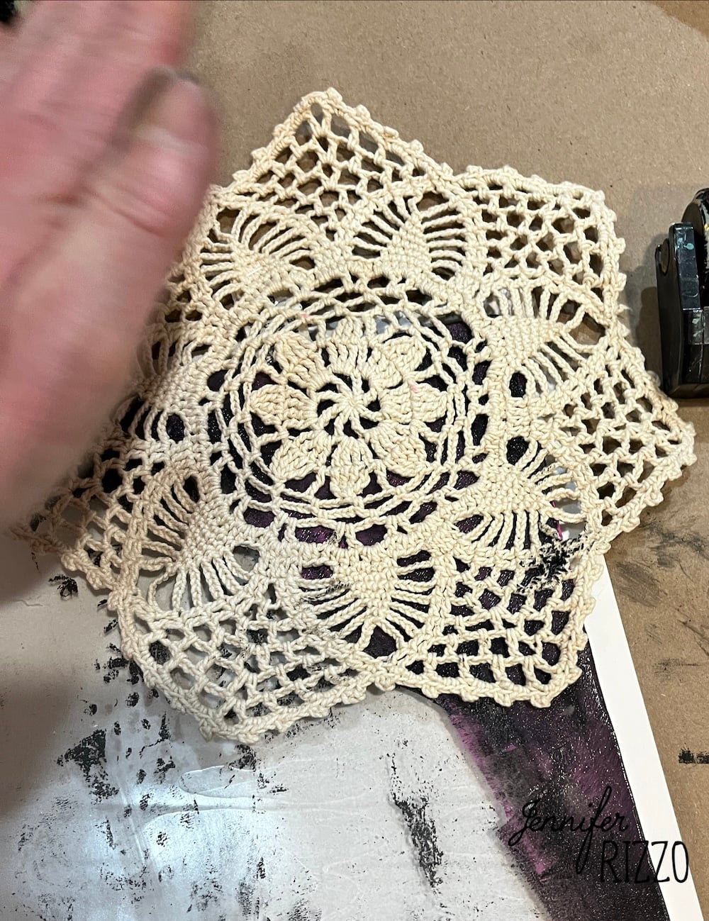 A hand holds a beige crochet doily on top of a piece of paper with printed text and a partially visible dark image.