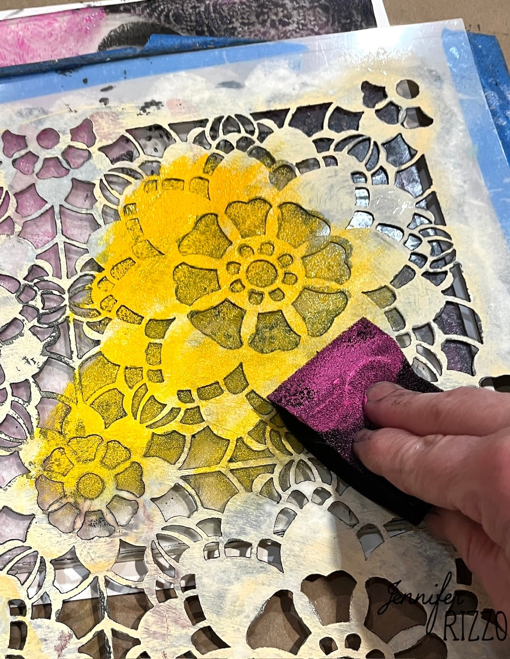 A hand uses a sponge to apply yellow paint over a floral-patterned stencil on paper.