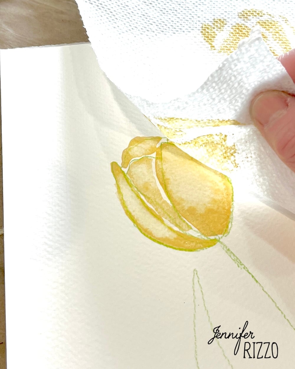 A hand uses a paper towel to blot a yellow watercolor tulip on white paper. The name "Jennifer Rizzo" appears in the bottom right corner.