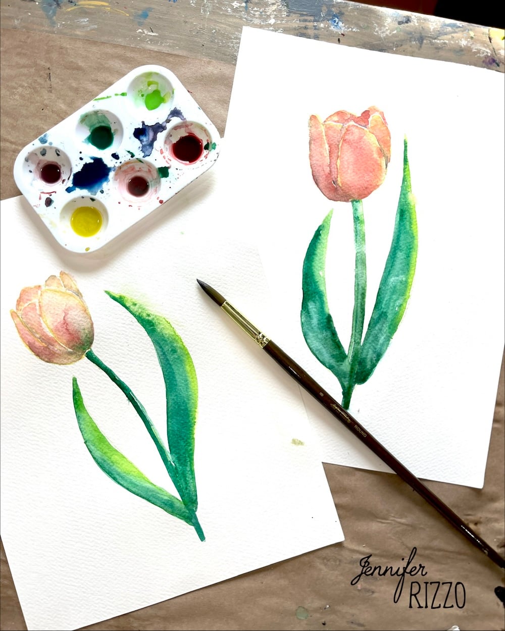 Two watercolor paintings of tulips on white paper beside a paint palette and a brush on a brown surface.
