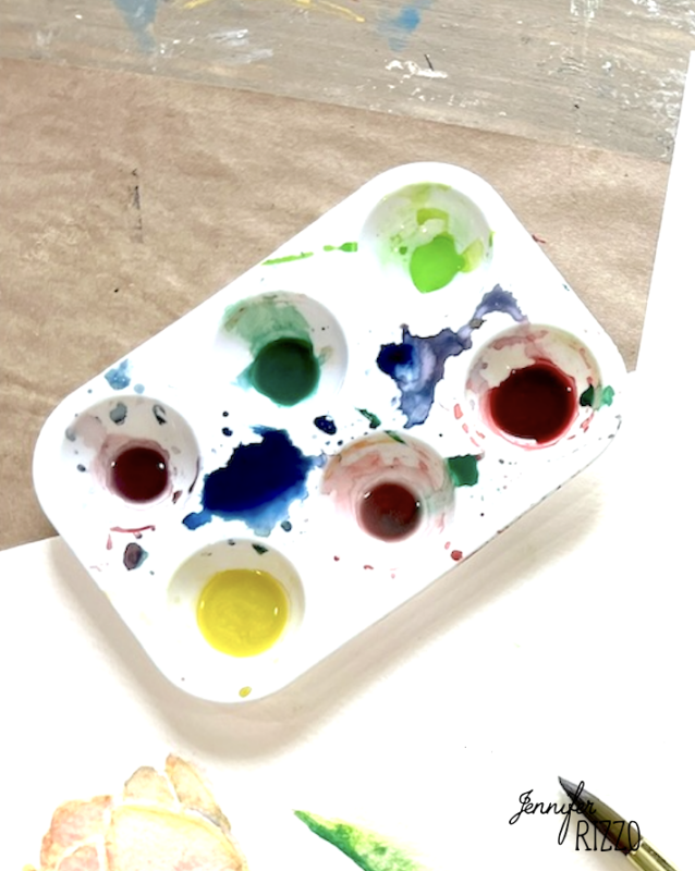 A plastic paint palette with six wells holds various colors including green, red, blue, and yellow, with paint splatters around the edges on a table.