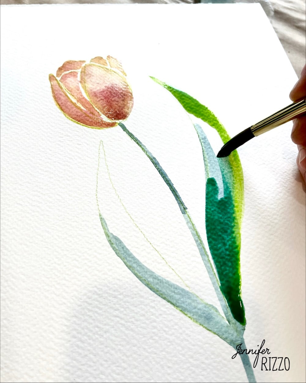 A hand uses a paintbrush to paint green leaves on a watercolor illustration of a tulip on textured white paper.