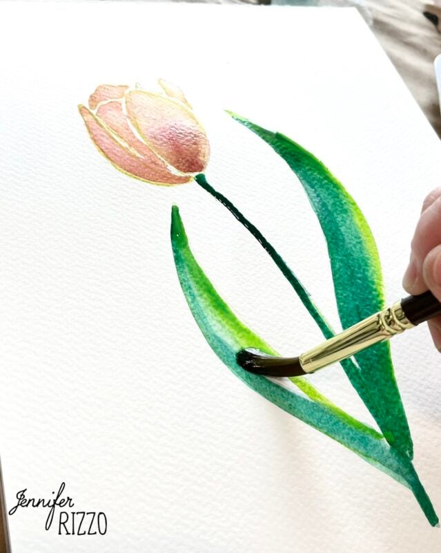 A hand uses a paintbrush to paint green leaves on a watercolor tulip on white paper. The name "Jennifer Rizzo" is printed in the lower left corner.