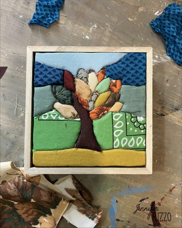 A framed Easy Kinusaiga Art piece depicts a tree with multicolored leaves on a green and yellow landscape, set against a blue and gray sky. Fabric scraps are scattered around.