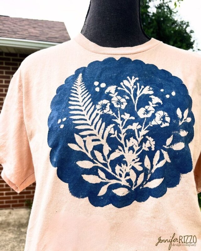 A light pink T-shirt displays a large blue circular design, created using Batik with Glue and Stencils, featuring white botanical silhouettes of ferns and flowers on the front.
