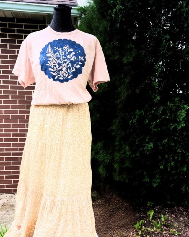 A mannequin displays a pink T-shirt with a blue floral print and a long, light yellow patterned skirt created using Batik with Glue and Stencils, set outdoors near a brick wall and greenery.