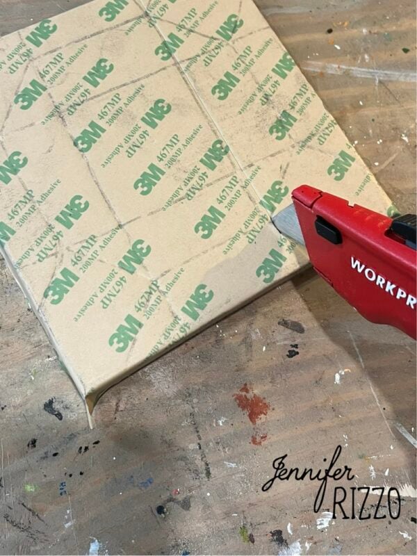 A red utility knife is used to cut excess adhesive-backed 3M paper from the edge of a wrapped rectangular board on a worktable, making Easy Kinusaiga Art projects simple and precise.