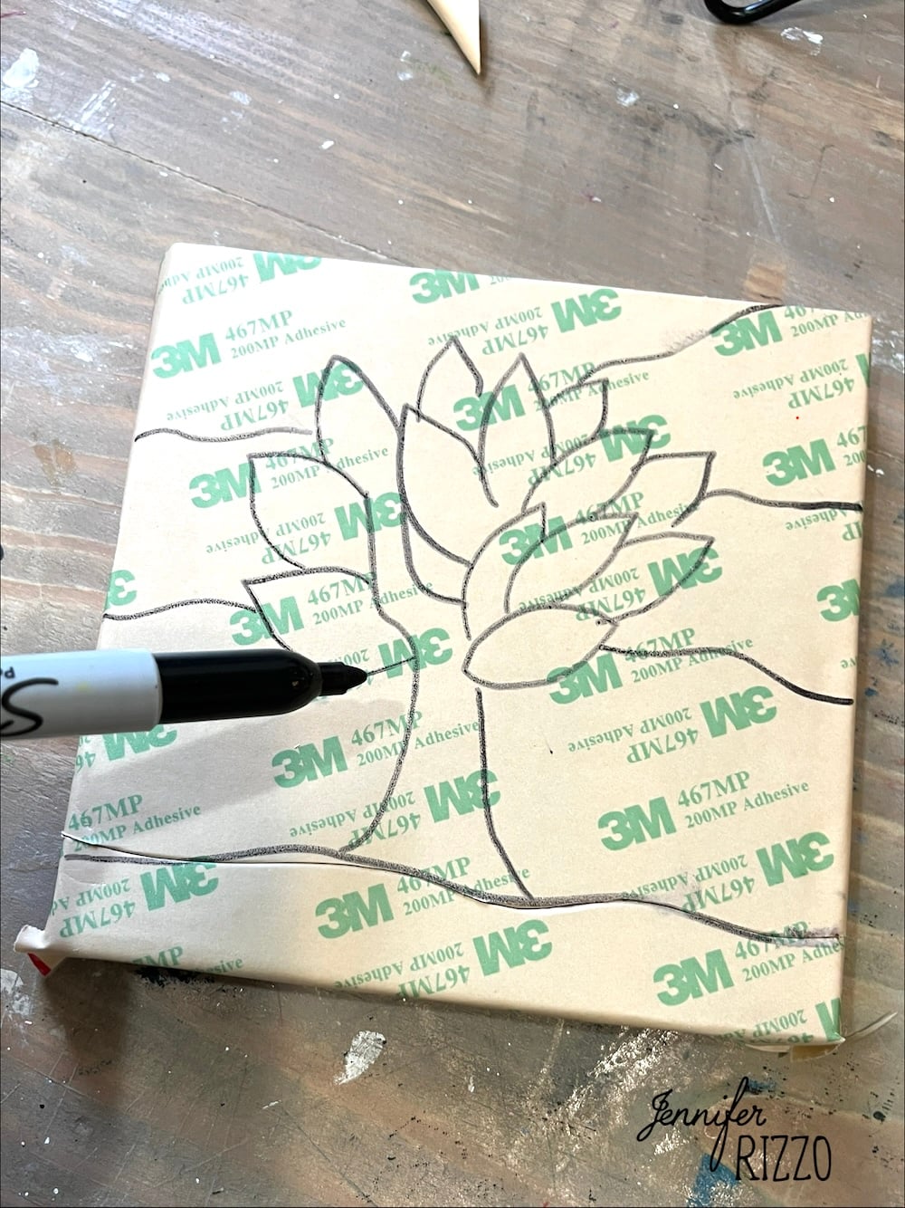 A hand draws a flower outline with a black marker on a piece of 3M adhesive sheet placed on a wooden surface, creating an Easy Kinusaiga Art design.