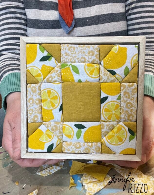 Hands holding a small framed fabric quilt block featuring yellow, white, and lemon-patterned fabrics arranged in a geometric design—an inspiring example of Easy Kinusaiga Art.
