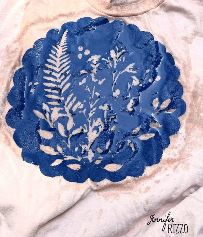 A white fabric features a circular blue Batik with Glue and Stencils design of leaves and ferns, partially wet, displaying the name "Jennifer Rizzo" in the bottom right corner.