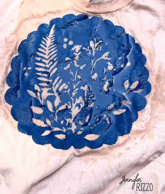 A white fabric features a circular blue Batik with Glue and Stencils design of leaves and ferns, partially wet, displaying the name "Jennifer Rizzo" in the bottom right corner.