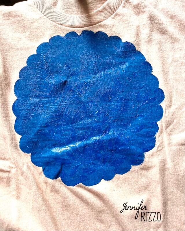 A white shirt featuring a large, textured, scalloped blue circle in the center—created using Batik with Glue and Stencils—and the text "Jennifer Rizzo" printed in black at the bottom right.