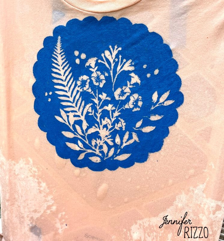 A beige fabric features a blue circular Batik with Glue and Stencils design, adorned with white floral and fern patterns, and the text "Jennifer Rizzo" in the bottom right corner.