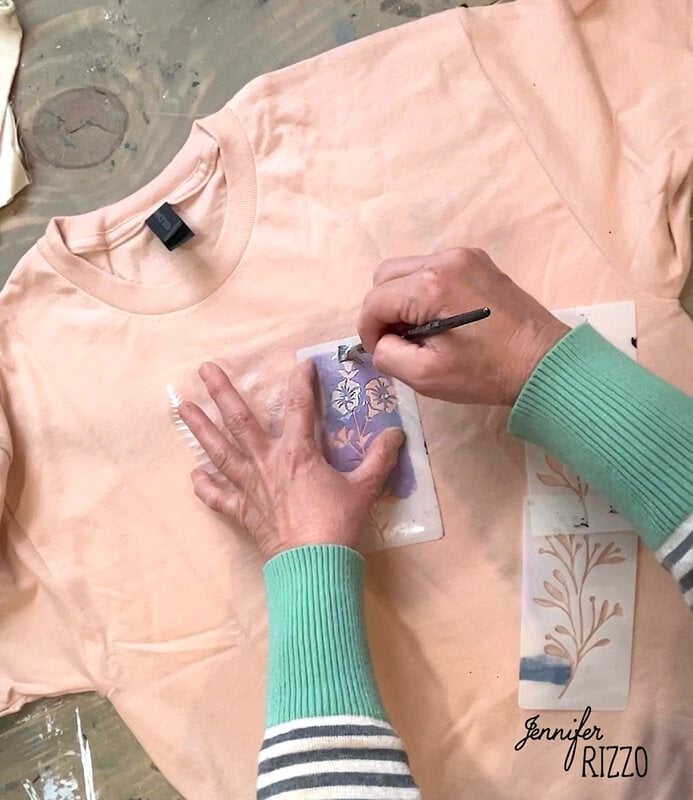 Person painting a floral stencil design in purple on a light pink t-shirt, using Batik with Glue and Stencils technique, with stencils and paint visible on the table.