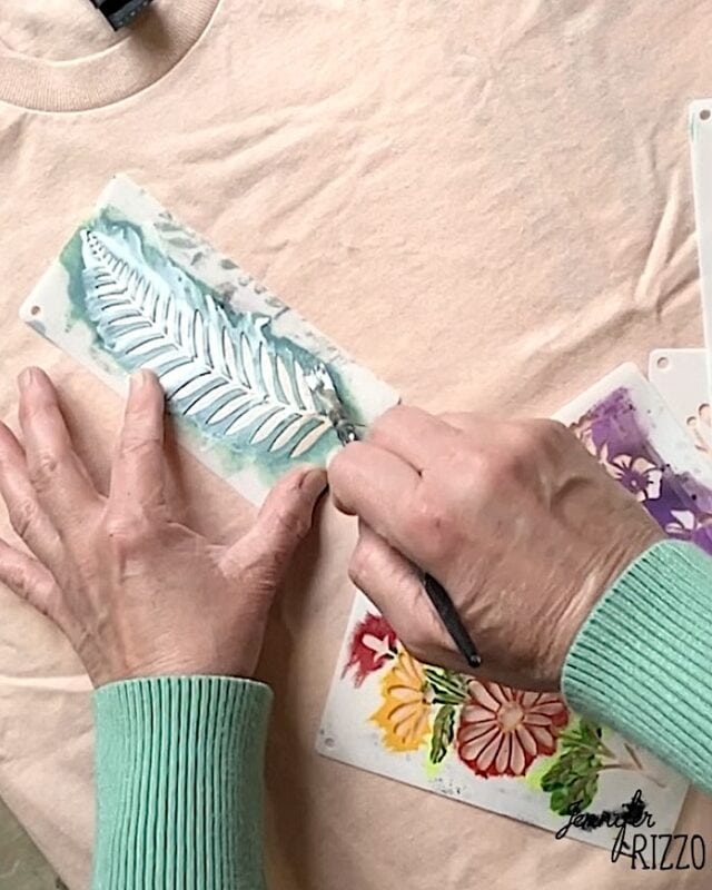 A person in a green sweater uses Batik with Glue and Stencils, applying a stencil and brush to paint a white leaf design on light-colored fabric.