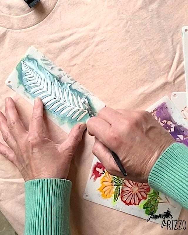 A person in a green sweater uses Batik with Glue and Stencils, applying a stencil and brush to paint a white leaf design on light-colored fabric.
