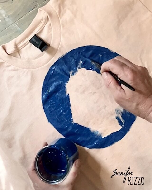 A person uses Batik with Glue and Stencils to paint a large blue circle on a light-colored T-shirt with a brush, holding a cup of blue paint. The name "Jennifer Rizzo" appears in the corner.