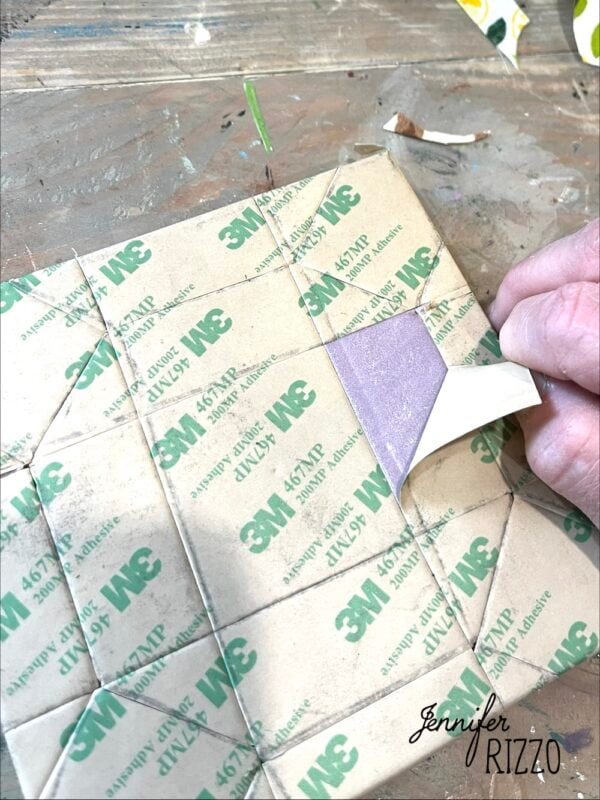 A hand peels back a piece of adhesive backing from a sheet of 3M 467MP transfer tape on a cardboard surface, perfect for Easy Kinusaiga Art projects. "Jennifer Rizzo" is written in the lower right corner.