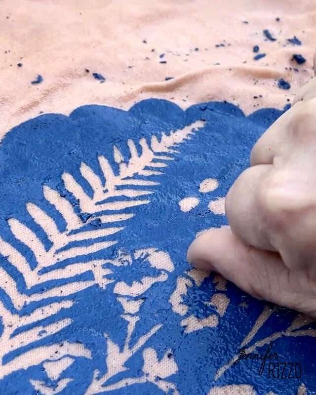 A hand applies blue textured paint over a fern leaf stencil on a peach-colored surface, creating an effect reminiscent of Batik with Glue and Stencils.