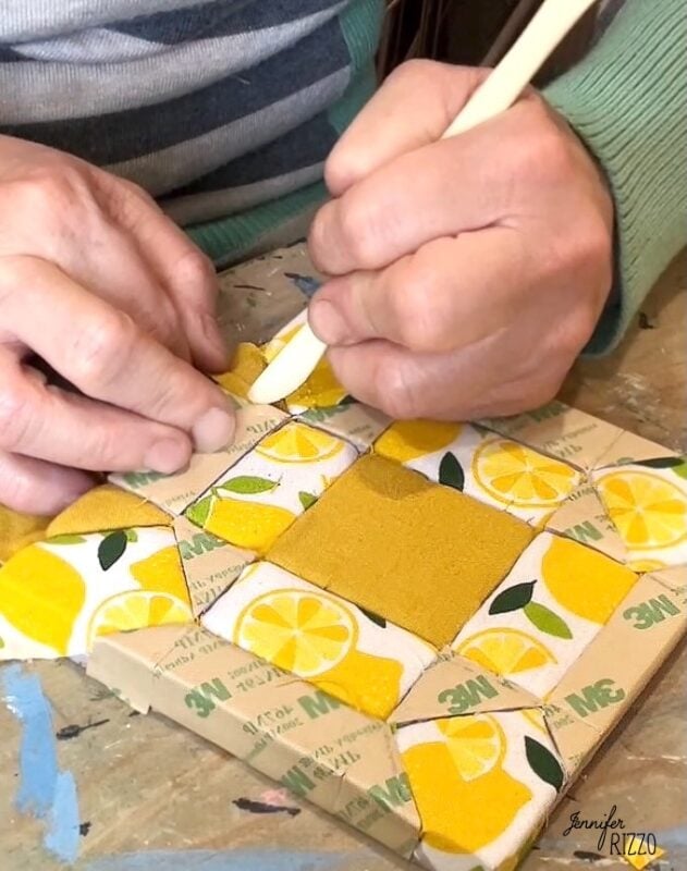 Hands using a wooden tool to smooth lemon-patterned fabric decoupage—an Easy Kinusaiga Art technique—on a surface with visible 3M adhesive backing.