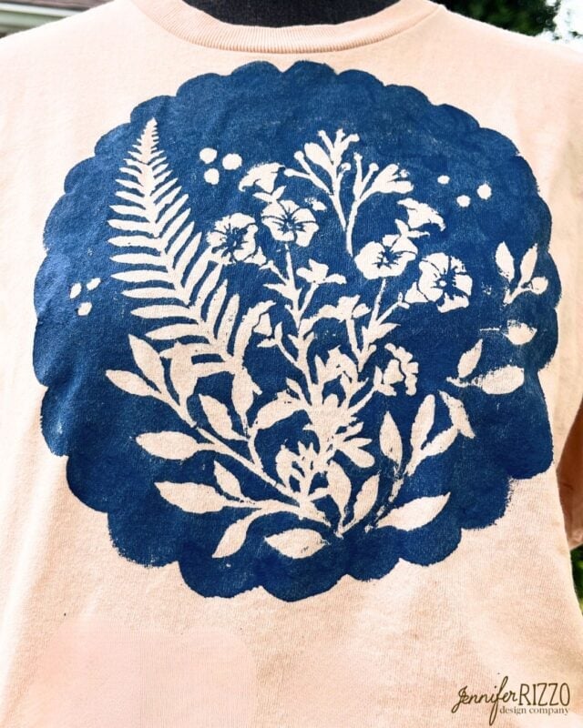 A light-colored shirt features a dark blue stencil design of leaves, ferns, and flowers inside a scalloped circular shape, inspired by Batik with Glue and Stencils. "jennifer RIZZO design company" is printed in the corner.