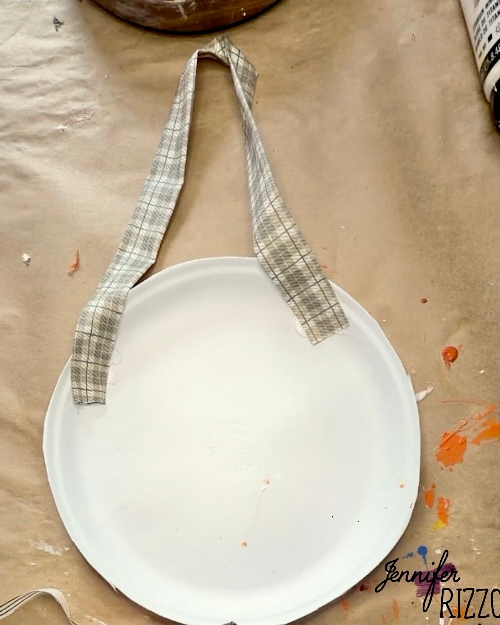 A plain white paper plate with a plaid fabric handle attached, placed on a craft table with paint splatters.