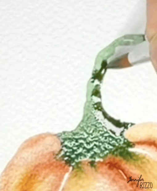A paintbrush colors the green stem of a watercolor pumpkin on textured paper.