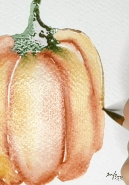 A hand paints the orange ridges of a watercolor pumpkin on white paper. The pumpkin has a green stem and soft blending of colors.