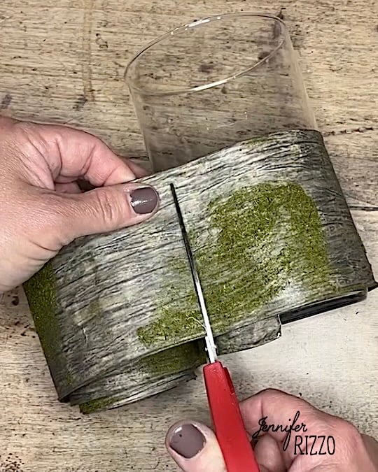 A person cuts a faux wood-textured decorative wrap with scissors next to a clear glass container on a wooden surface.